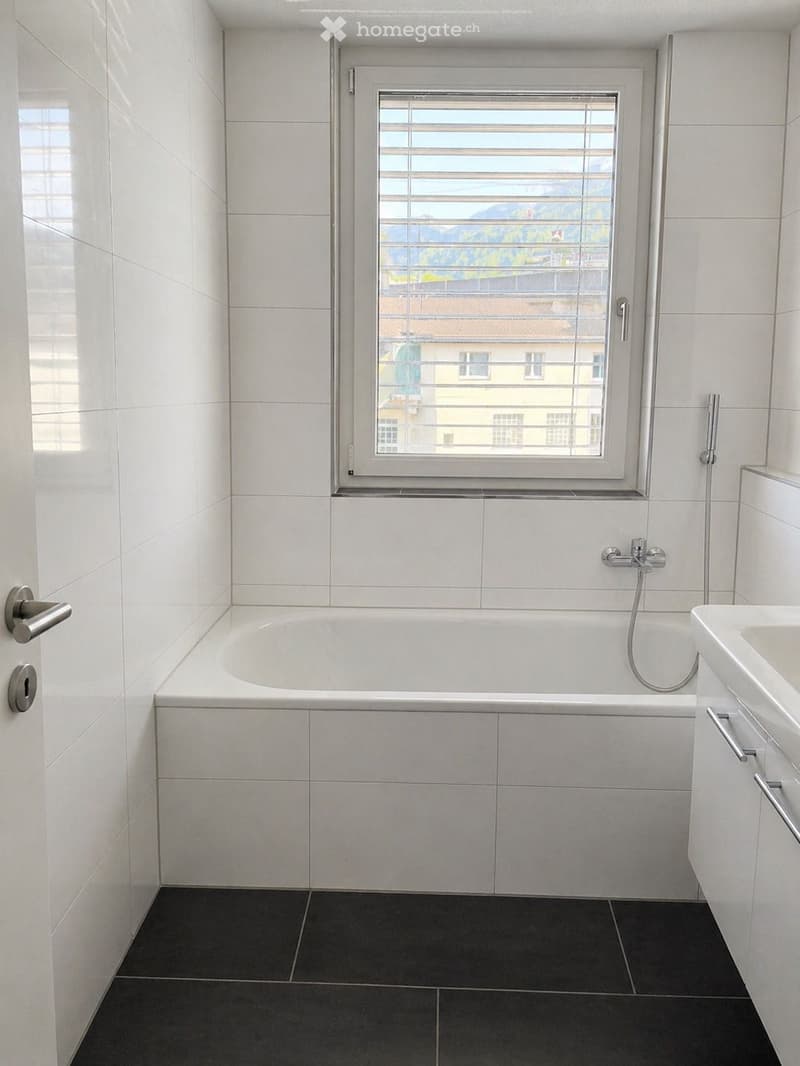 Modern bathroom with white tiled walls, a bathtub, a sink with a cabinet, and a window offering a scenic view.
