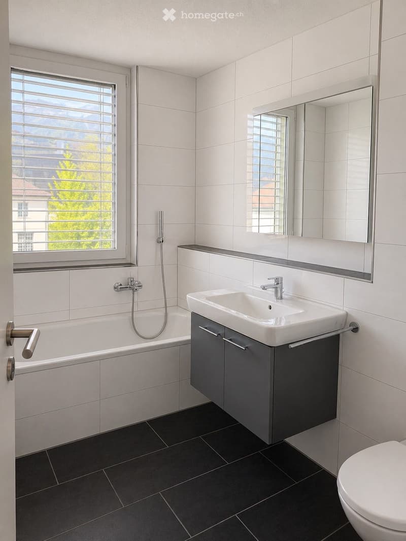 Modern bathroom with white tiles, a bathtub, a sink with a cabinet, a large mirror, and a toilet. The room features a window with blinds and a sleek, contemporary design.