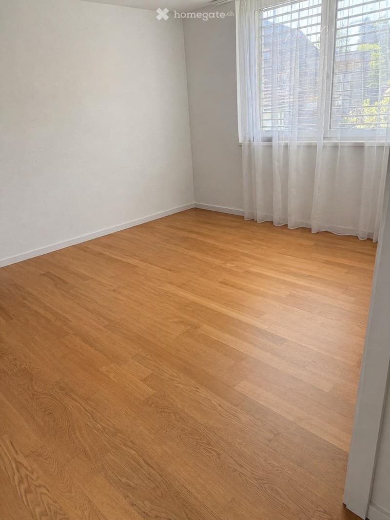 Empty room with light wooden flooring and white walls, featuring a window with sheer curtains.