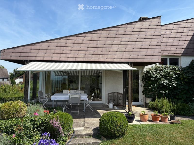 House with brown roof, white walls, patio with white table and chairs under striped awning, green garden with various plants and shrubs