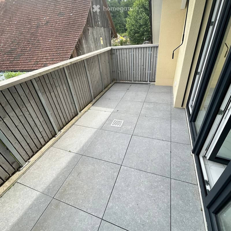 Balcony with grey tiled floor, wooden railing, and a view of neighboring building and greenery.