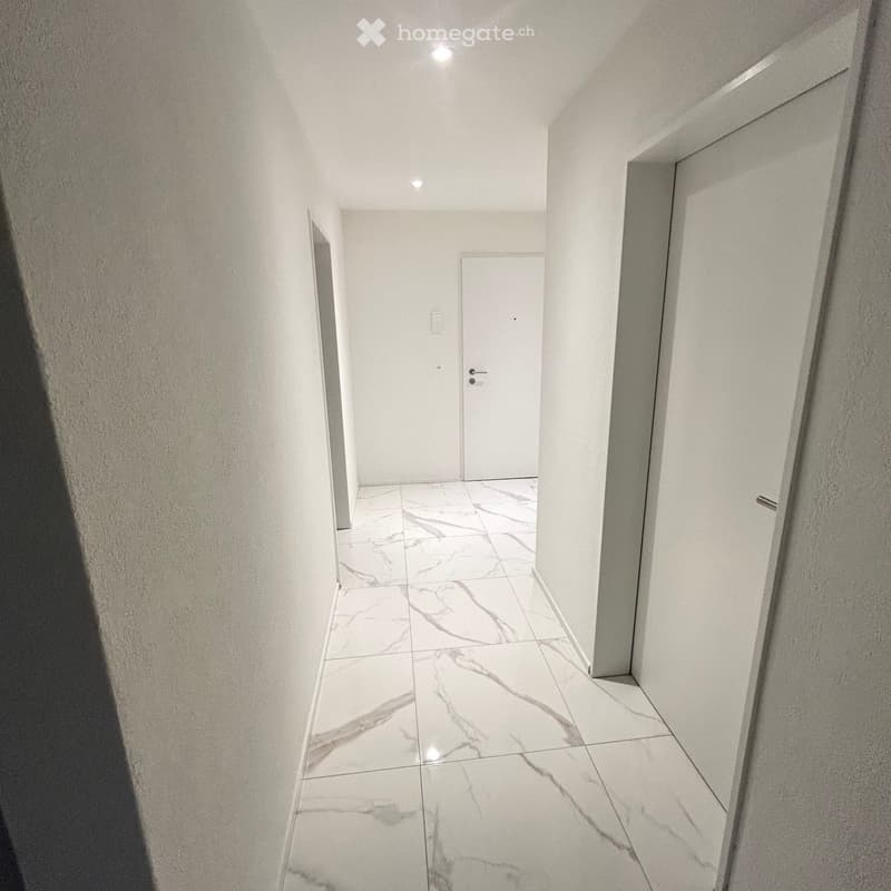 Clean, modern hallway with white marble tiled flooring and white walls. Features recessed ceiling lighting and two closed doors.
