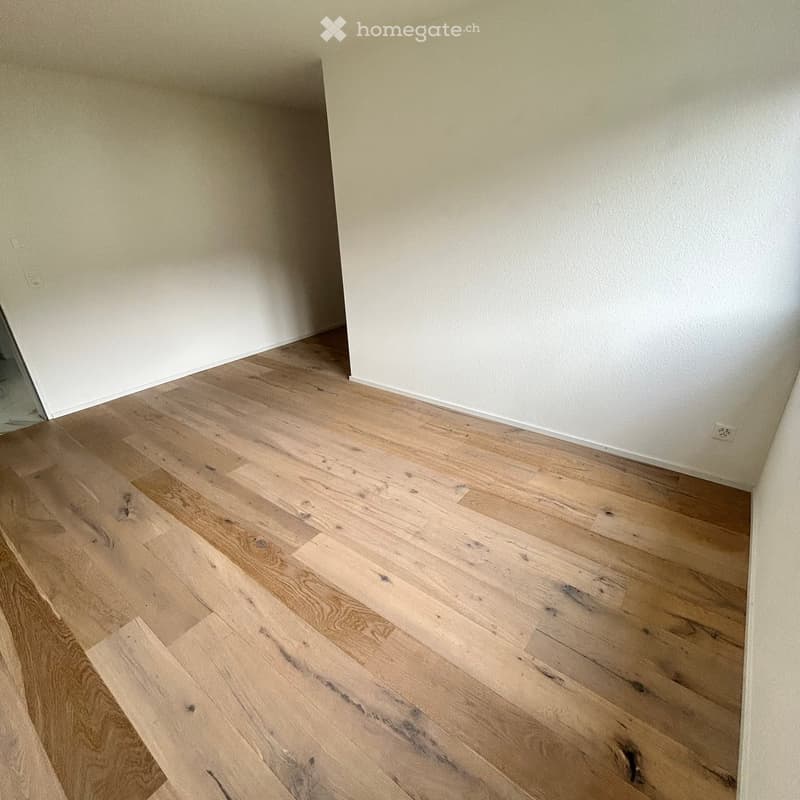 Empty room with light wooden flooring and white walls, showing a corner view with visible electrical outlets.