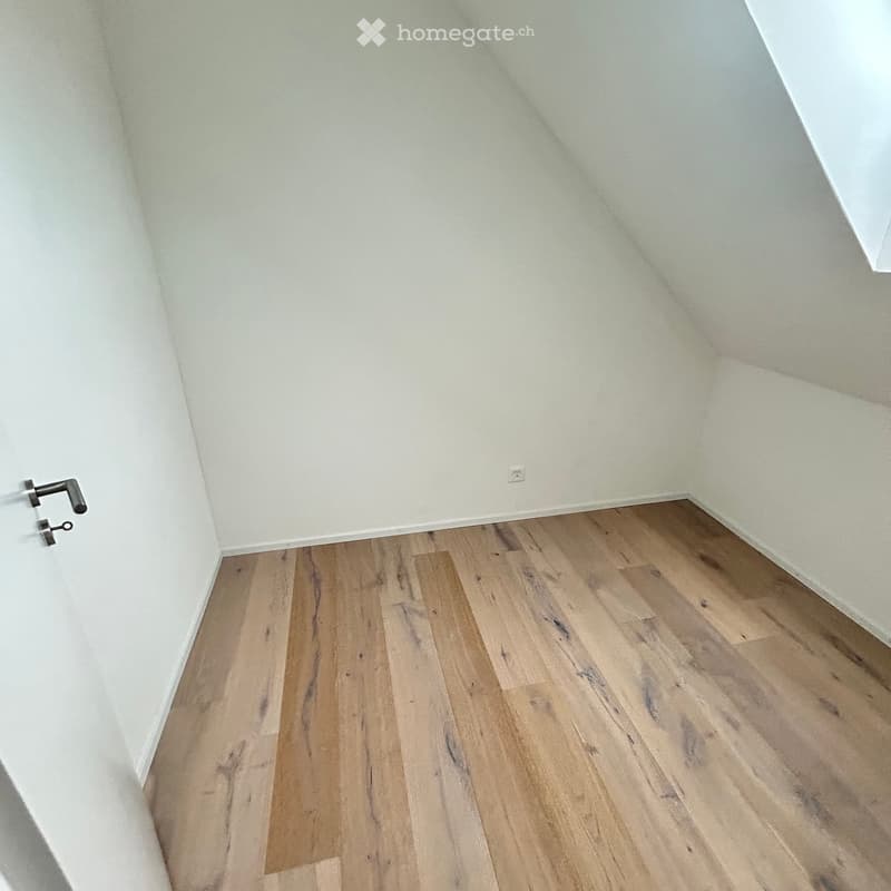 Empty room with light wooden flooring and white walls, featuring a door handle on the left side.