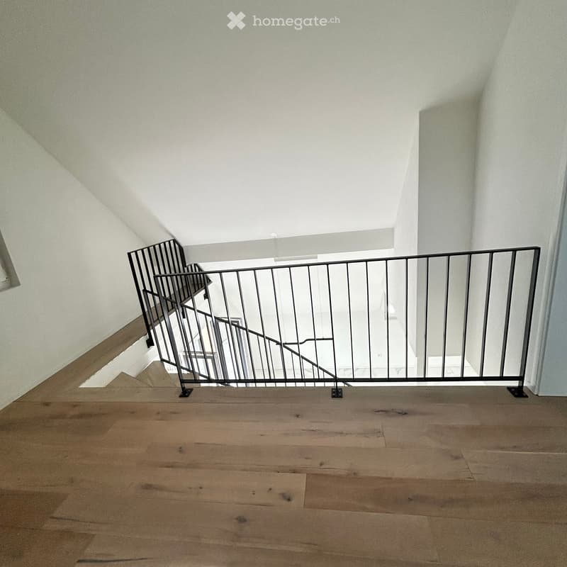 Empty hallway with wooden flooring and a metal railing leading to an upper level. The walls are white and the ceiling is sloped, allowing natural light to enter.