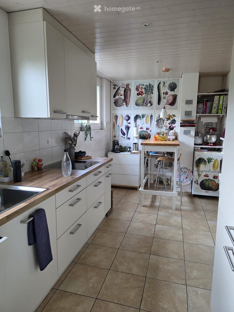 Modern kitchen with white cabinets, wooden countertops, and tiled floor. Features a central island with stools, a sink, stove, and various kitchen appliances.