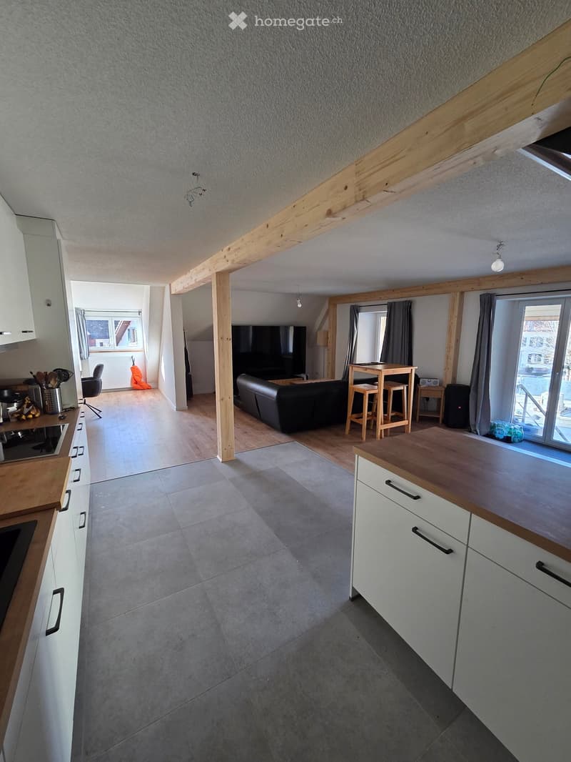 Open-plan living and dining area with a sofa, dining table with stools, kitchen cabinets, and a stove. The space features wooden beams on the ceiling and large windows with views.