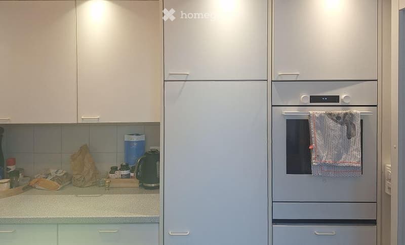 Modern kitchen with white cabinets, a built-in oven, and a refrigerator. The counter has various items including a kettle, bottles, and a bag.