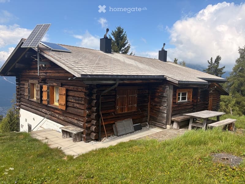 A wooden log cabin with a grey shingled roof, solar panels, and a small outdoor seating area with a picnic table and benches.