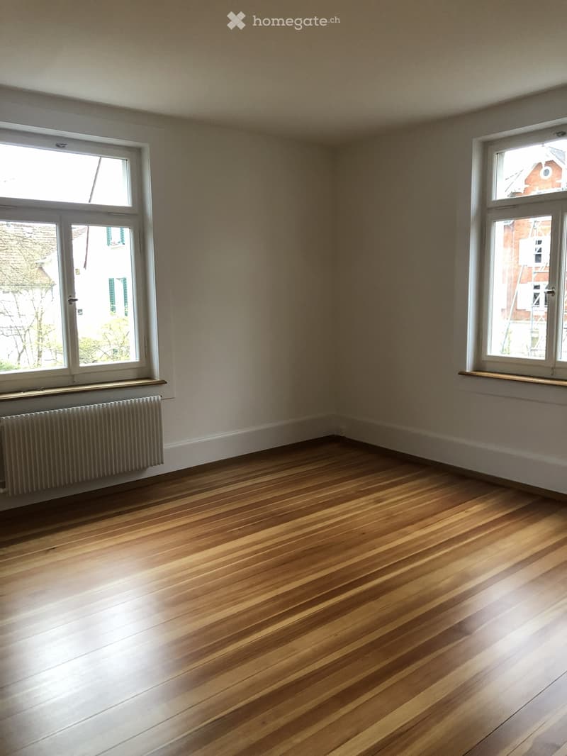 A view of a hallway with wooden flooring, white walls, and multiple windows allowing natural light to enter. A radiator is visible on the right side.