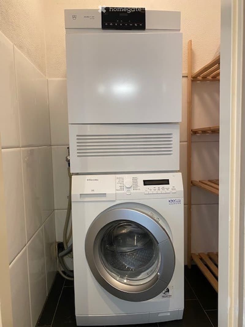 A compact bathroom with a stacked washing machine and dishwasher unit, white tiled walls, and a wooden shelf.
