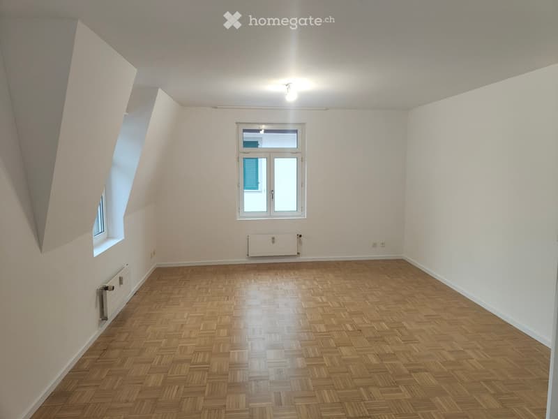 Empty room with wooden herringbone flooring, white walls, and two windows, one with blue shutters visible through the glass.