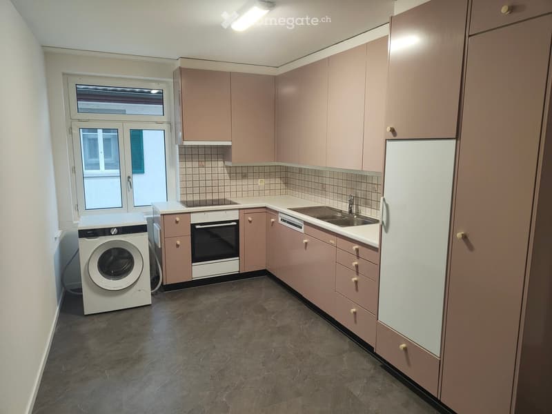 Modern kitchen with light pink cabinets, stainless steel appliances, and a large window. Includes a refrigerator, oven, dishwasher, sink, and washing machine.