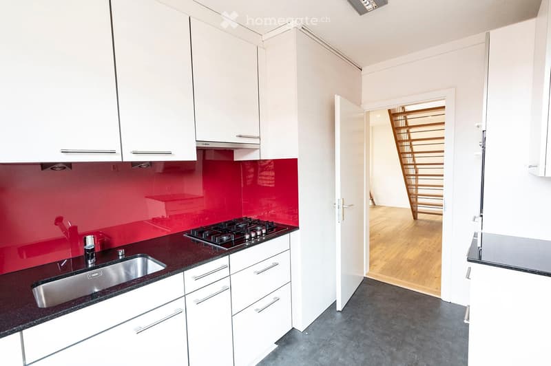 Modern kitchen with white cabinets, dark countertops, stainless steel sink, and a red backsplash. A wooden staircase is visible in the adjacent room.