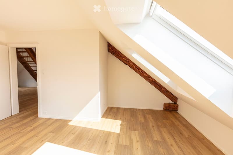 Empty attic room with wooden flooring, white walls, and skylight windows allowing natural light to enter. A wooden beam runs diagonally across the space, and a doorway leads to a staircase.