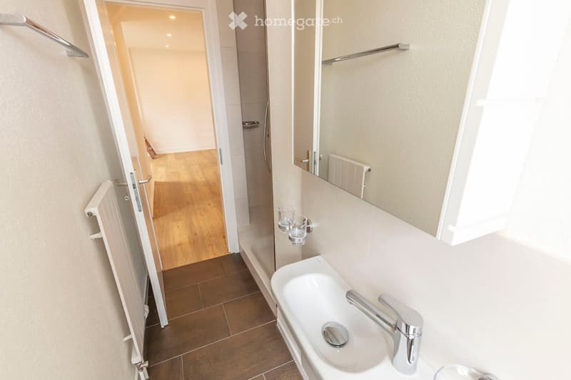 Modern bathroom with a white sink, chrome faucet, large mirror, radiator, and two cups on a shelf. The floor is tiled, and the walls are painted in a light neutral color. An open doorway leads to another room with wooden flooring.