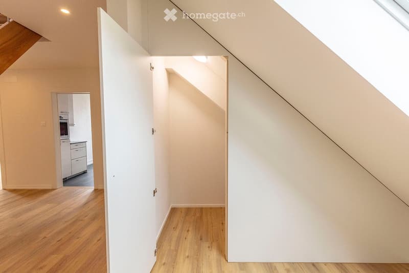 A narrow, well-lit hallway with wooden flooring and white walls, featuring an open door leading to a kitchen area with cabinets and appliances.
