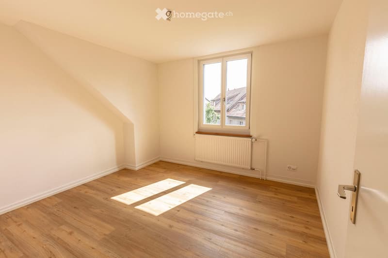 Empty room with wooden flooring, white walls, a window with a radiator below, and a door with a golden handle.