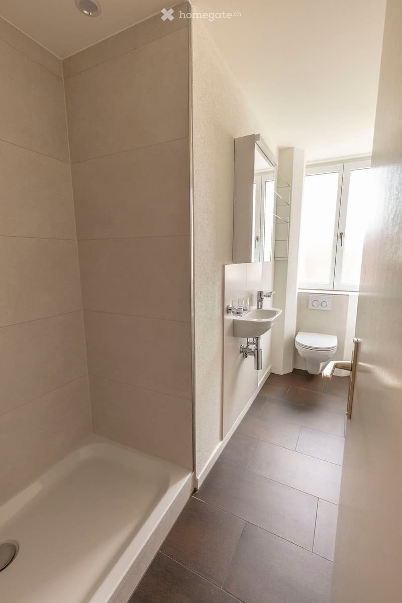 Modern bathroom with a bathtub, sink, toilet, and mirror. Light-colored tiles and a window provide natural light.