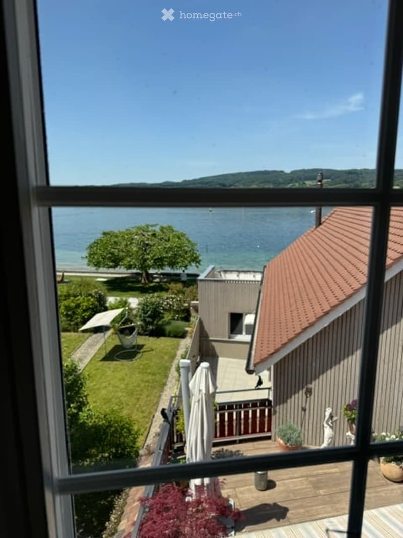 View from a balcony overlooking a serene lake with a tree, lawn, and nearby buildings.