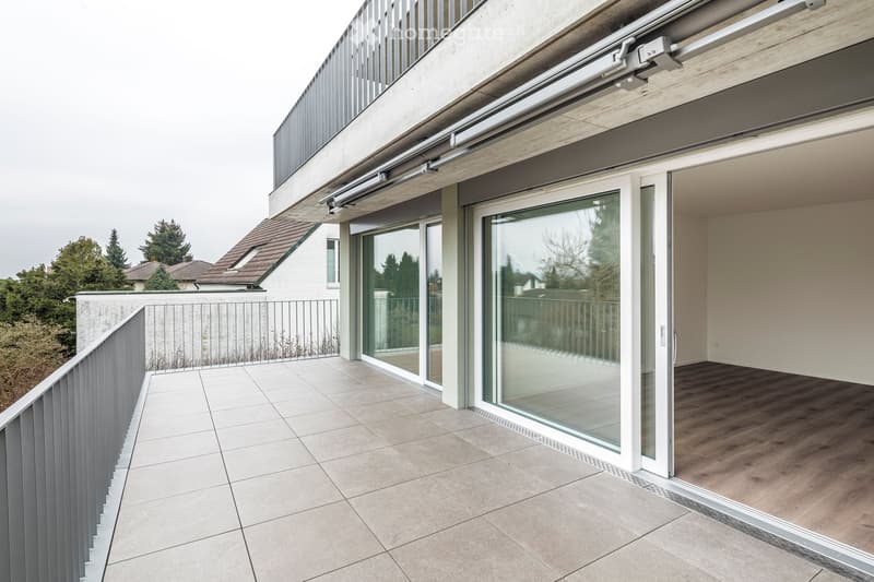 Modern balcony with large glass sliding doors, tiled floor, metal railing, and a view of the surrounding greenery.