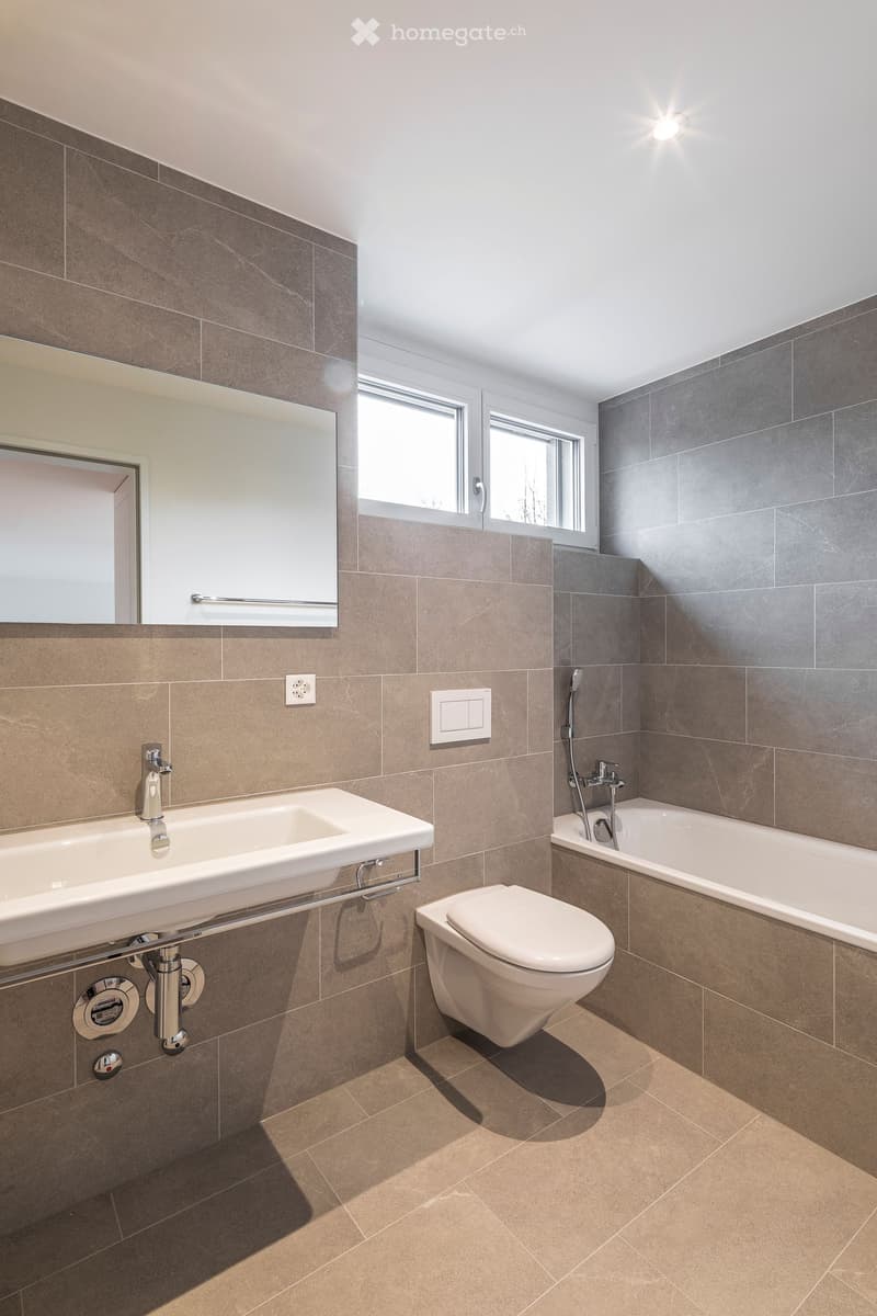 Modern bathroom with grey tiled walls, a white sink, toilet, bathtub, and a large mirror above the sink. Natural light from a window illuminates the space.