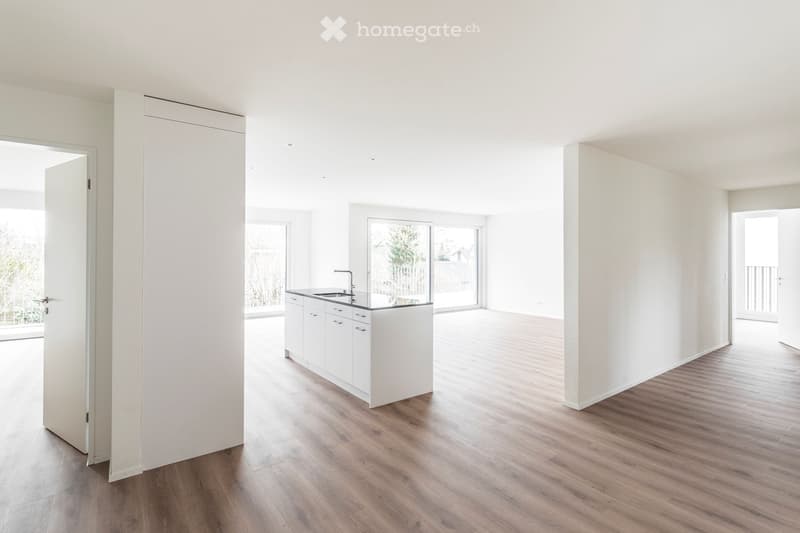 Open-plan unfurnished room with light-colored walls, wooden flooring, and a small white kitchen area with cabinets and a sink.
