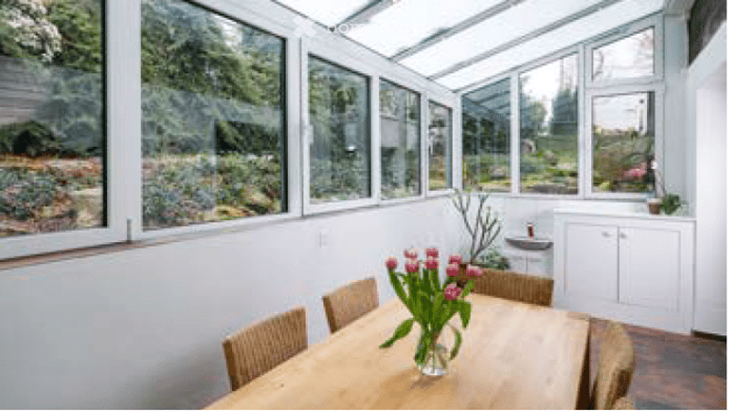 Bright conservatory with large windows offering a view of the garden, featuring a wooden dining table with chairs and a vase of pink tulips, a small sink, and white cabinetry.