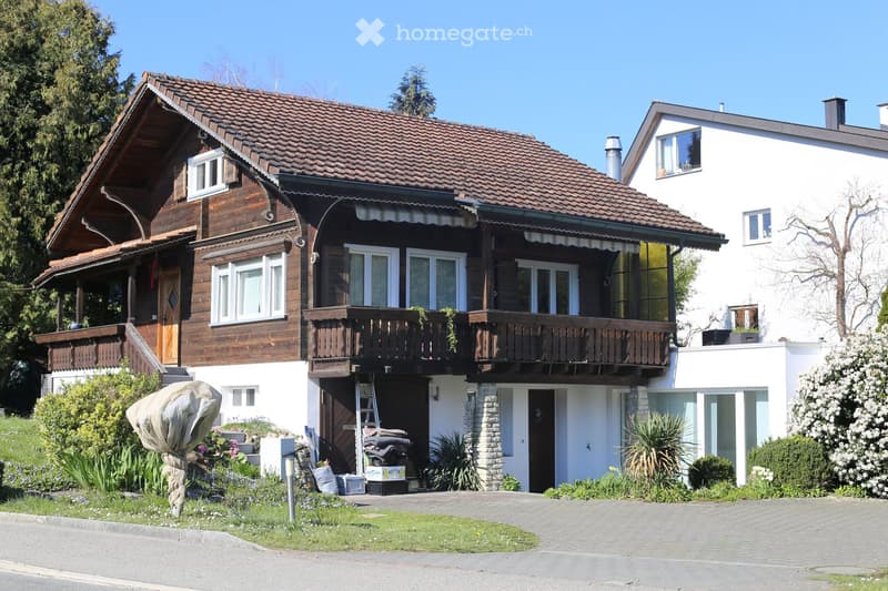 Two-story house with a wooden balcony, tiled roof, and modern white facade. The house features multiple windows, a covered entrance, and a well-maintained garden with various plants and shrubs.