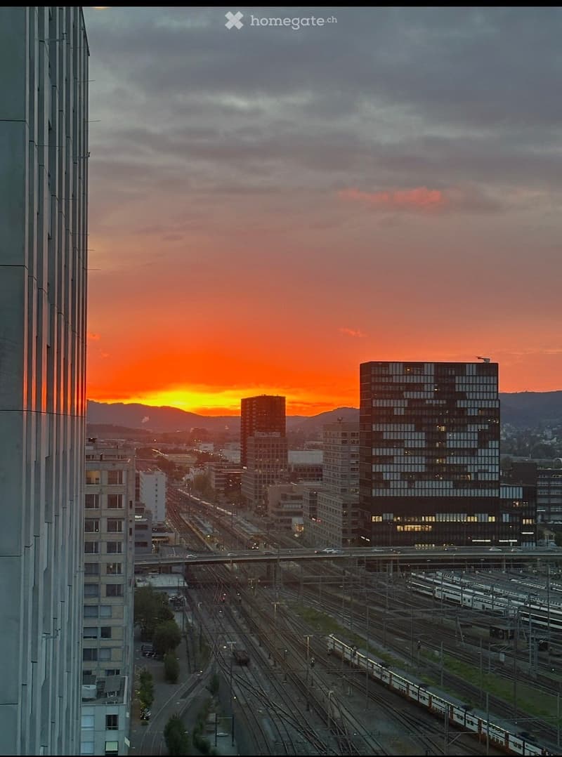 A panoramic view of a cityscape at sunset, featuring a train station with multiple tracks and trains, surrounded by modern buildings and a vibrant orange sky.