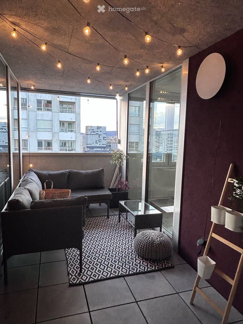 Modern balcony with dark grey sofa, coffee table, round stool, patterned rug, and potted plants. Large glass sliding doors open to a city view.