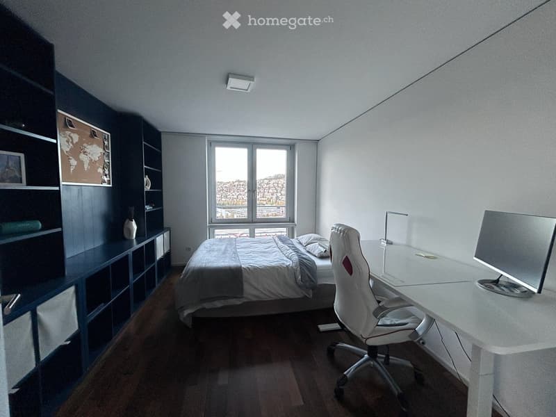 Modern bedroom with a bed, desk, chair, and large window offering a city view. Features a dark blue shelving unit with decorative items and a world map.