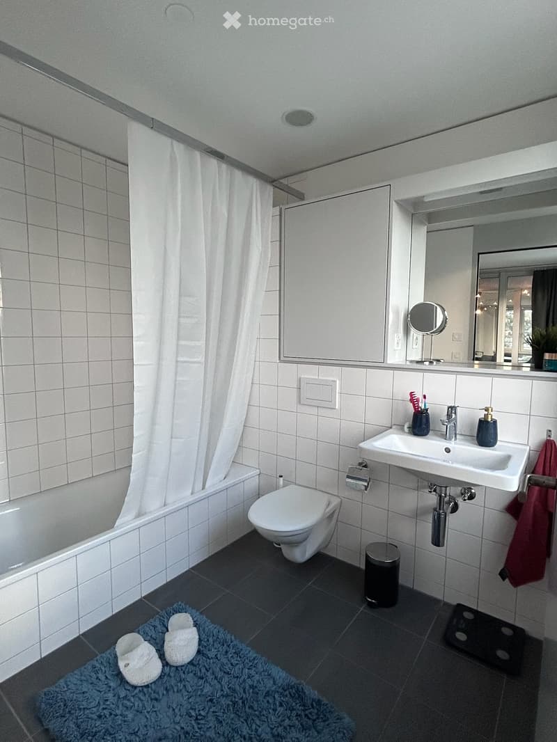 Modern bathroom with white tiled walls, a bathtub, toilet, sink, mirror, and a blue bath mat with slippers on the floor.