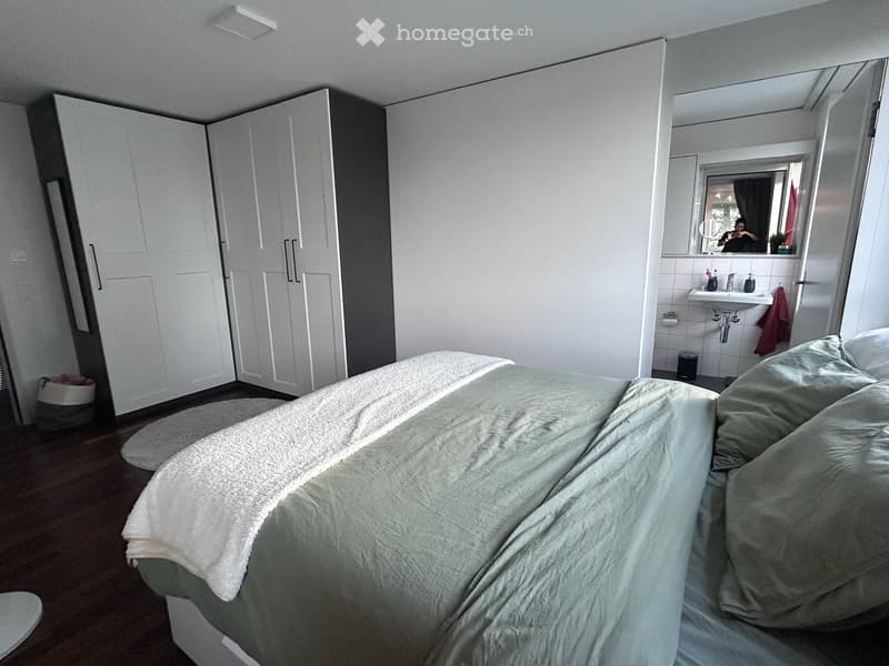 A modern bedroom with a neatly made bed, a large wardrobe, and an en-suite bathroom area featuring a sink, mirror, and towel.