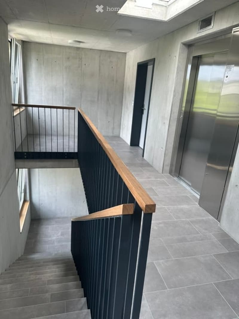 Modern hallway with concrete walls, tiled floor, staircase with wooden handrail, elevator, and a black door.