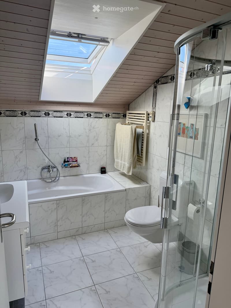 Modern bathroom with marble tiles, a bathtub, a toilet, a glass-enclosed shower, a towel rack with towels, and a skylight window.