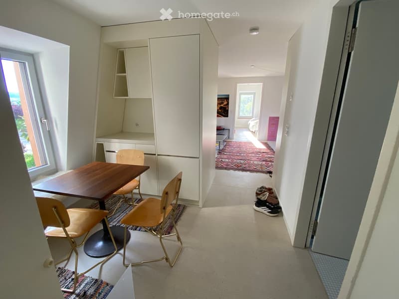 A small, modern furnished room with a wooden table, four chairs, and a built-in cabinet. The room has a view of another room with a bed and a colorful rug.
