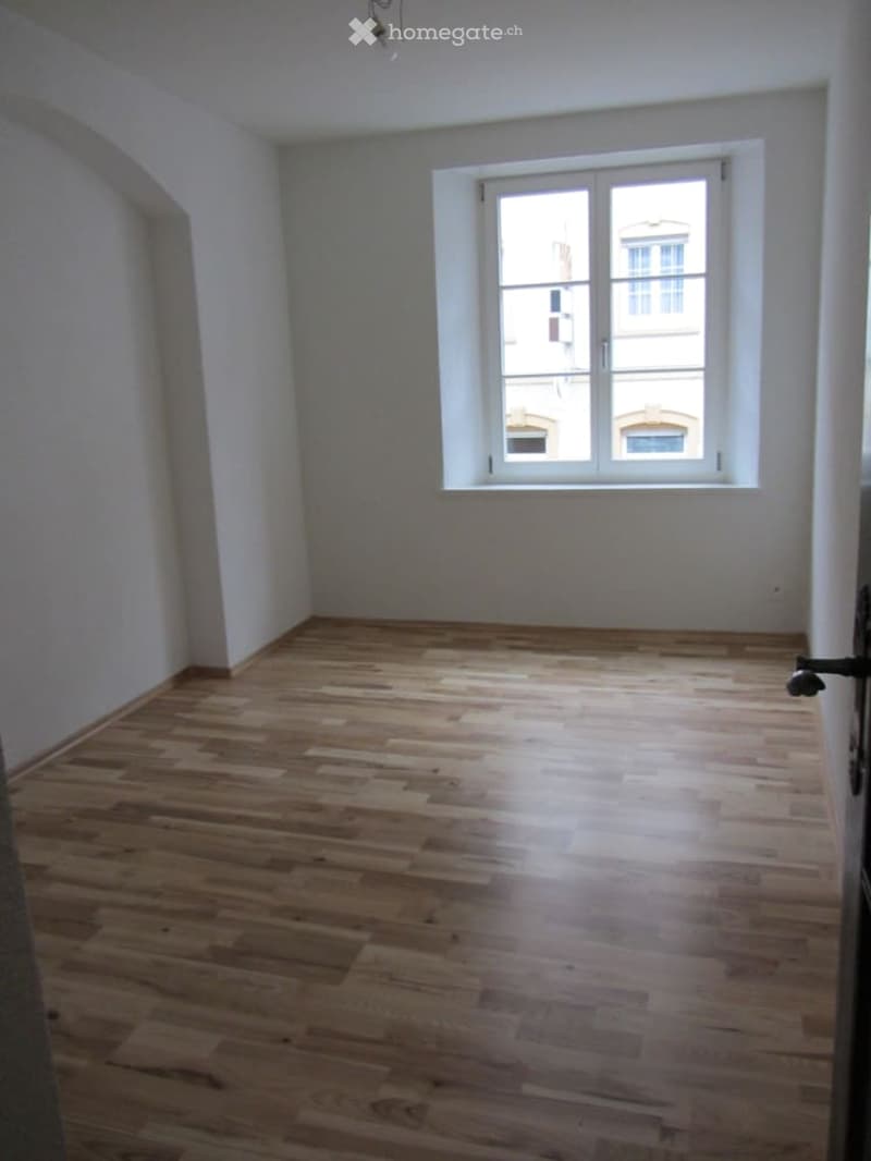 Completely empty room with wooden flooring and white walls, featuring a single window with white frame and glass panes.