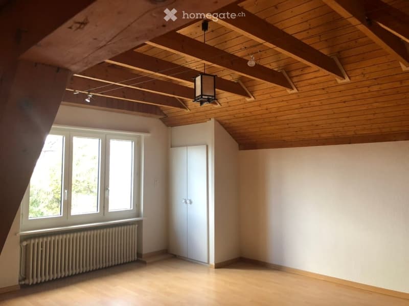 Empty attic room with wooden ceiling beams, exposed wooden ceiling, white walls, large windows, radiator, and built-in closet.