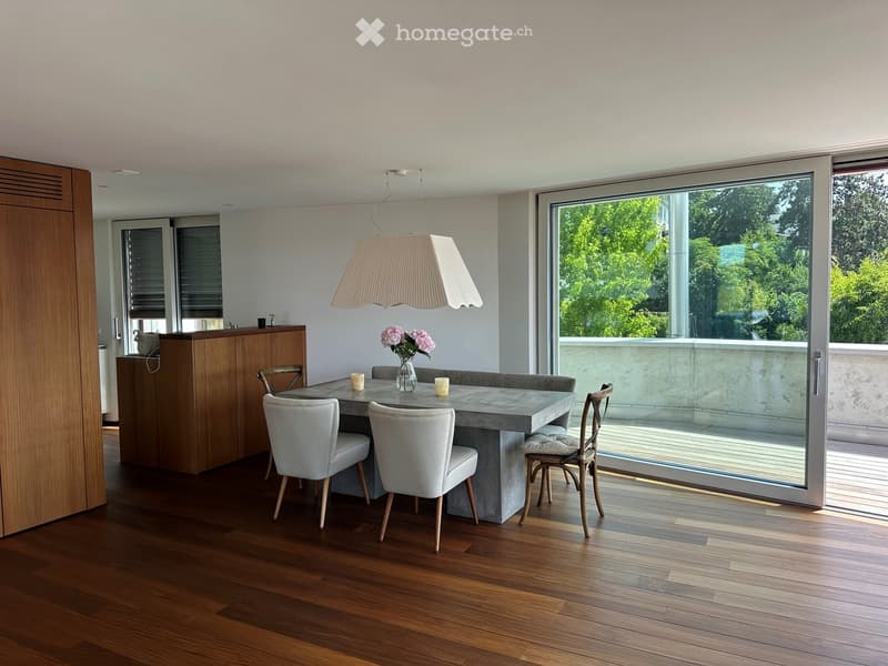 Modern dining area with a concrete table, four chairs, a hanging lamp, and a wooden cabinet. Large windows offer a view of greenery outside.
