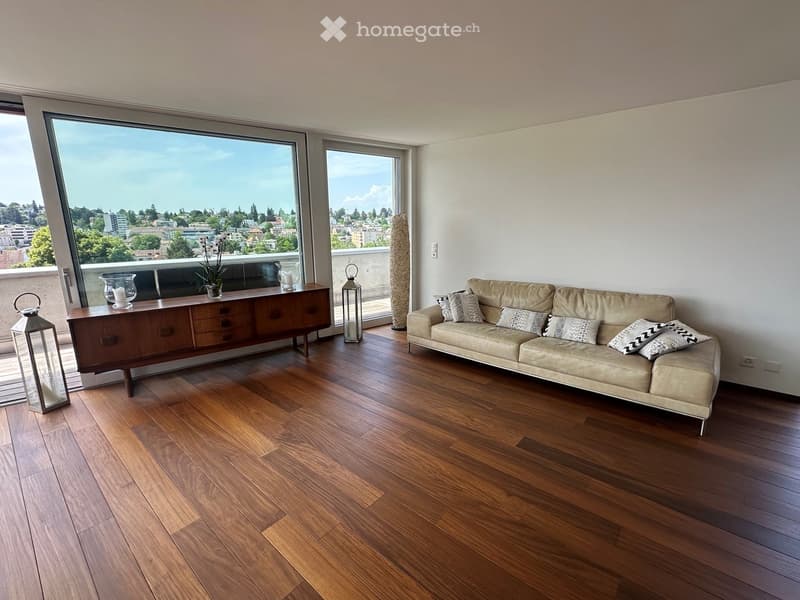 Modern living room with hardwood floors, large windows offering a city view, a beige sofa with patterned pillows, a wooden sideboard, and decorative lanterns.