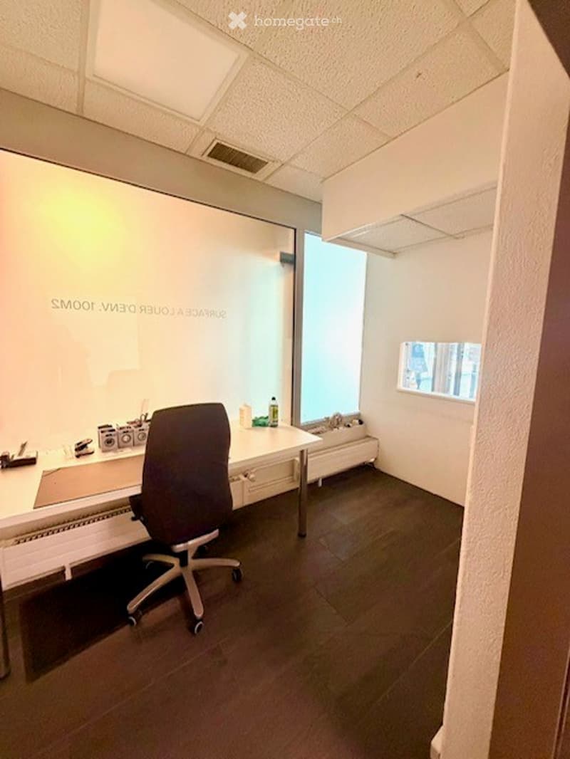 Modern office space with a white desk, ergonomic chair, large screen, laptop, and a bottle on the desk. The room has a polished dark floor and white walls.