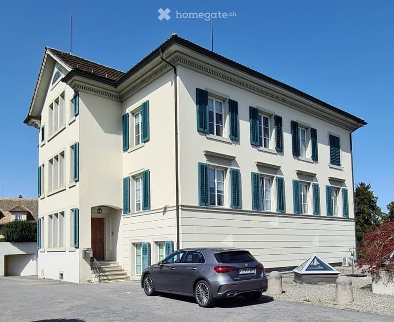 3-story building with a classical architectural style, featuring a white facade, black shutters, and a tiled roof. There is a car parked in the front of the building.