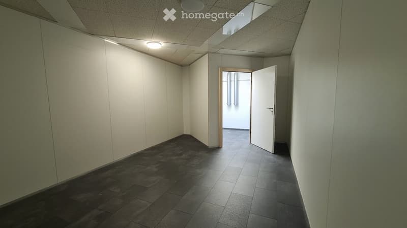 The image shows a spacious office space with gray walls, a tiled floor, and recessed lighting in the ceiling. The room has a plain, minimalist design with a white door leading to another area. The space appears to be empty and ready for use.