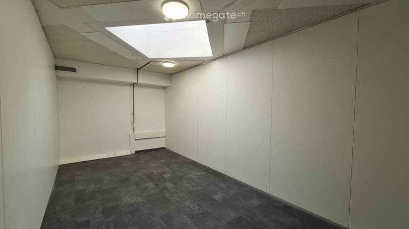 The image shows a spacious office space with white walls, a tiled gray floor, and recessed lighting in the ceiling. The room appears to be empty, with no furniture or other objects visible.
