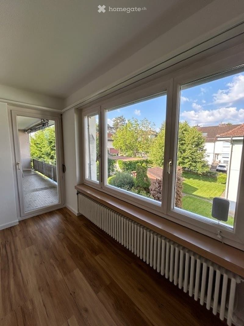 Bright, empty room with wooden flooring, large windows offering a view of a green garden, and a balcony accessible through a glass door.