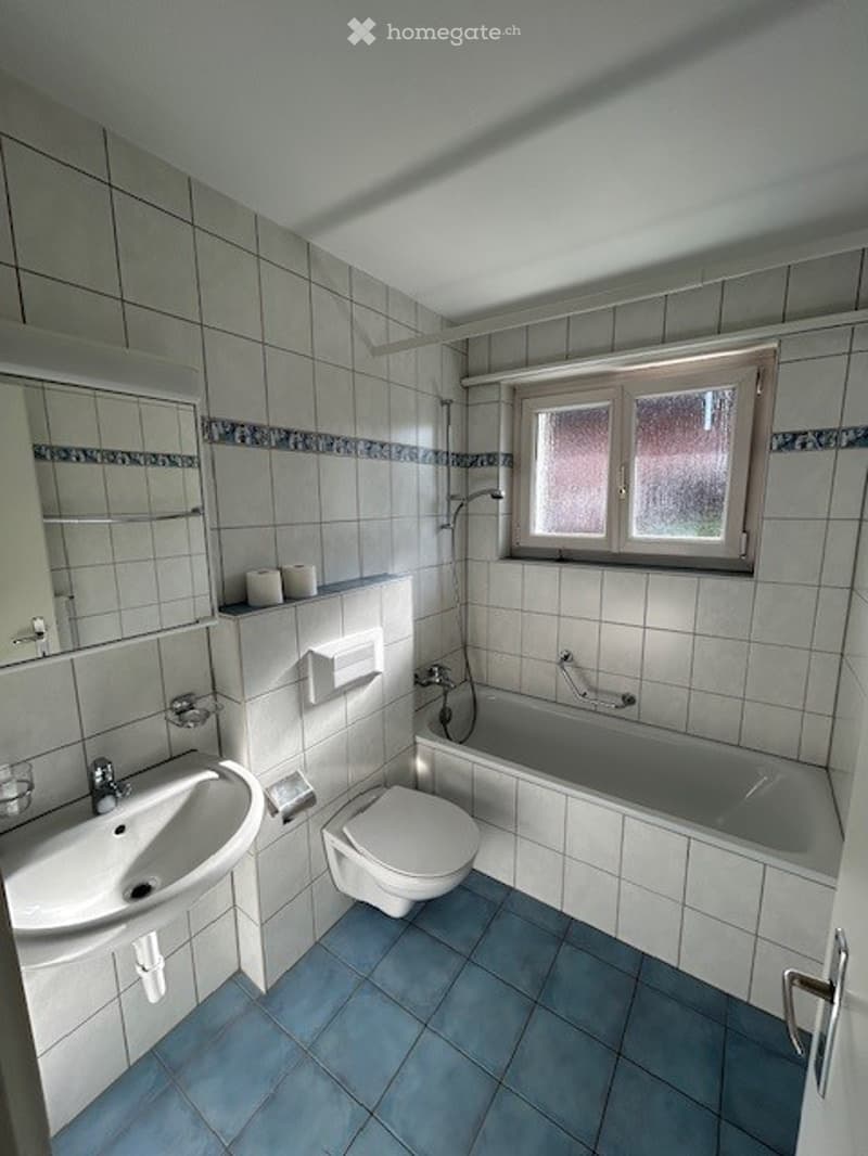 Bathroom with white tiles, blue floor tiles, a sink, toilet, bathtub, and a window with frosted glass.