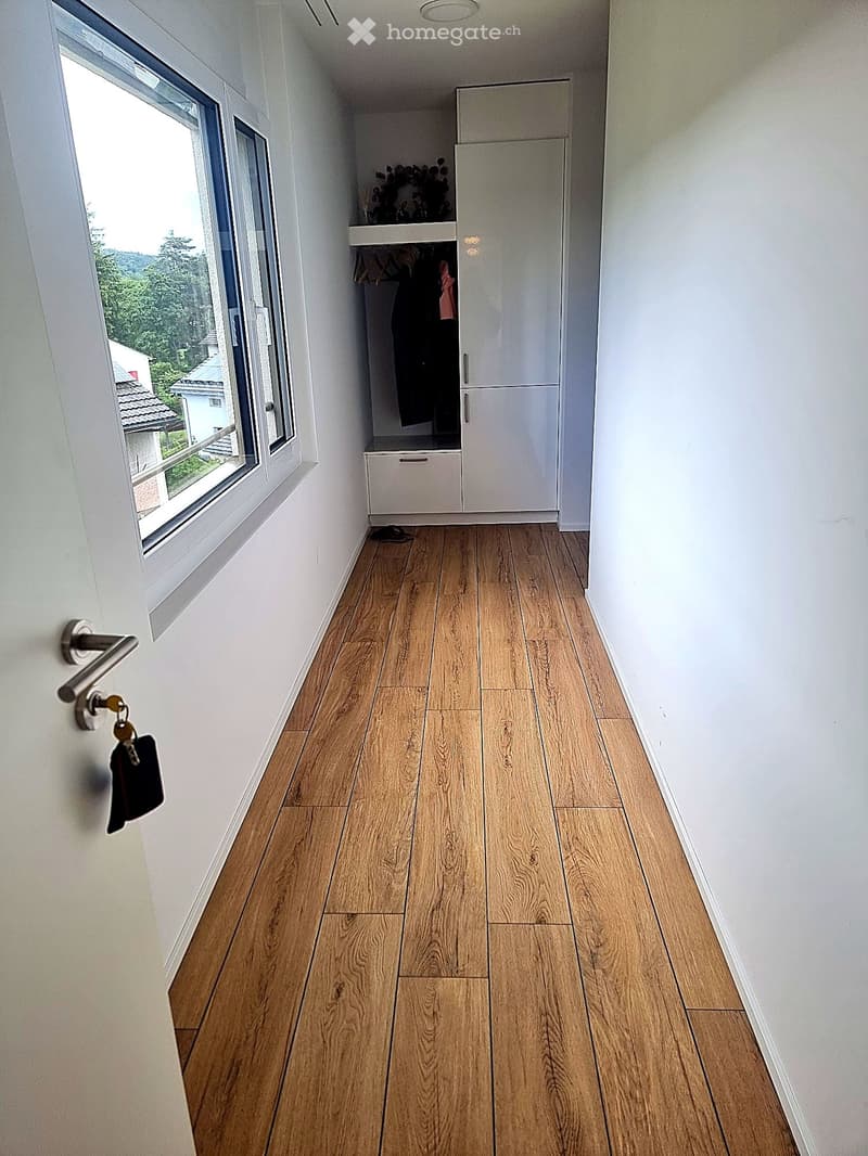 A narrow hallway with light wooden flooring, white walls, and a built-in white wardrobe with hanging space and a small drawer. A large window on the left side offers a view of greenery outside.