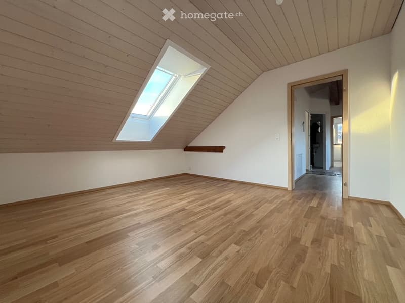 Empty attic room with wooden flooring, white walls, and a skylight window. The room features a slanted wooden ceiling and a small built-in shelf.