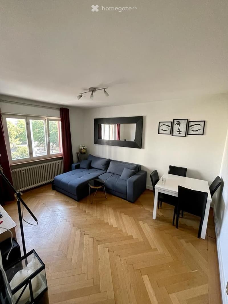 Modern living room with wooden flooring, large window, blue sectional sofa, small coffee table, dining table with chairs, and wall-mounted mirror.